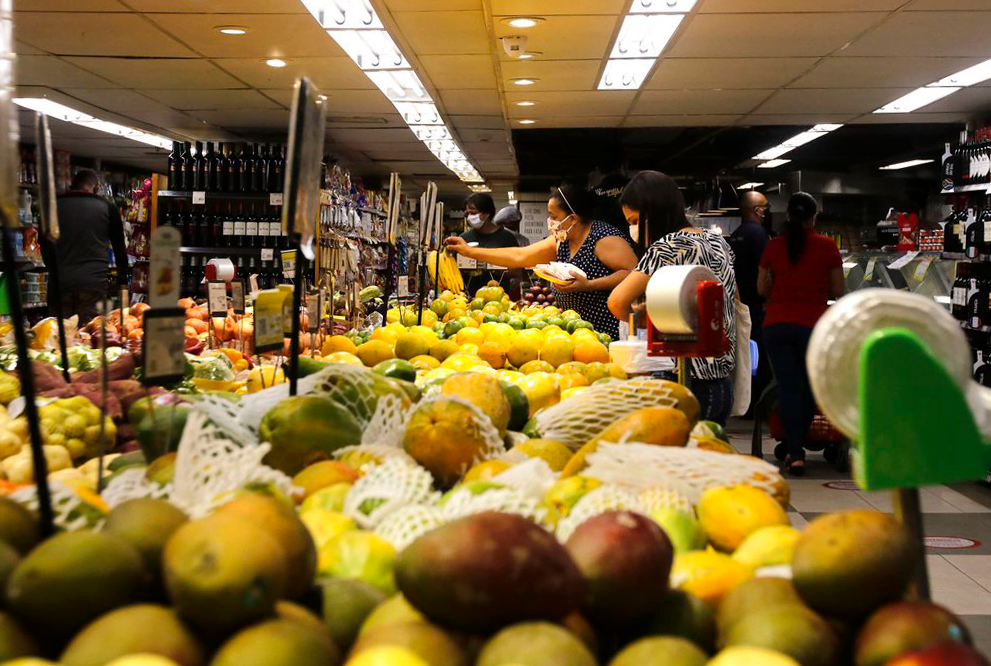 Foto de frutas em supermercado. No fundo, pessoas de máscara fazem compras.