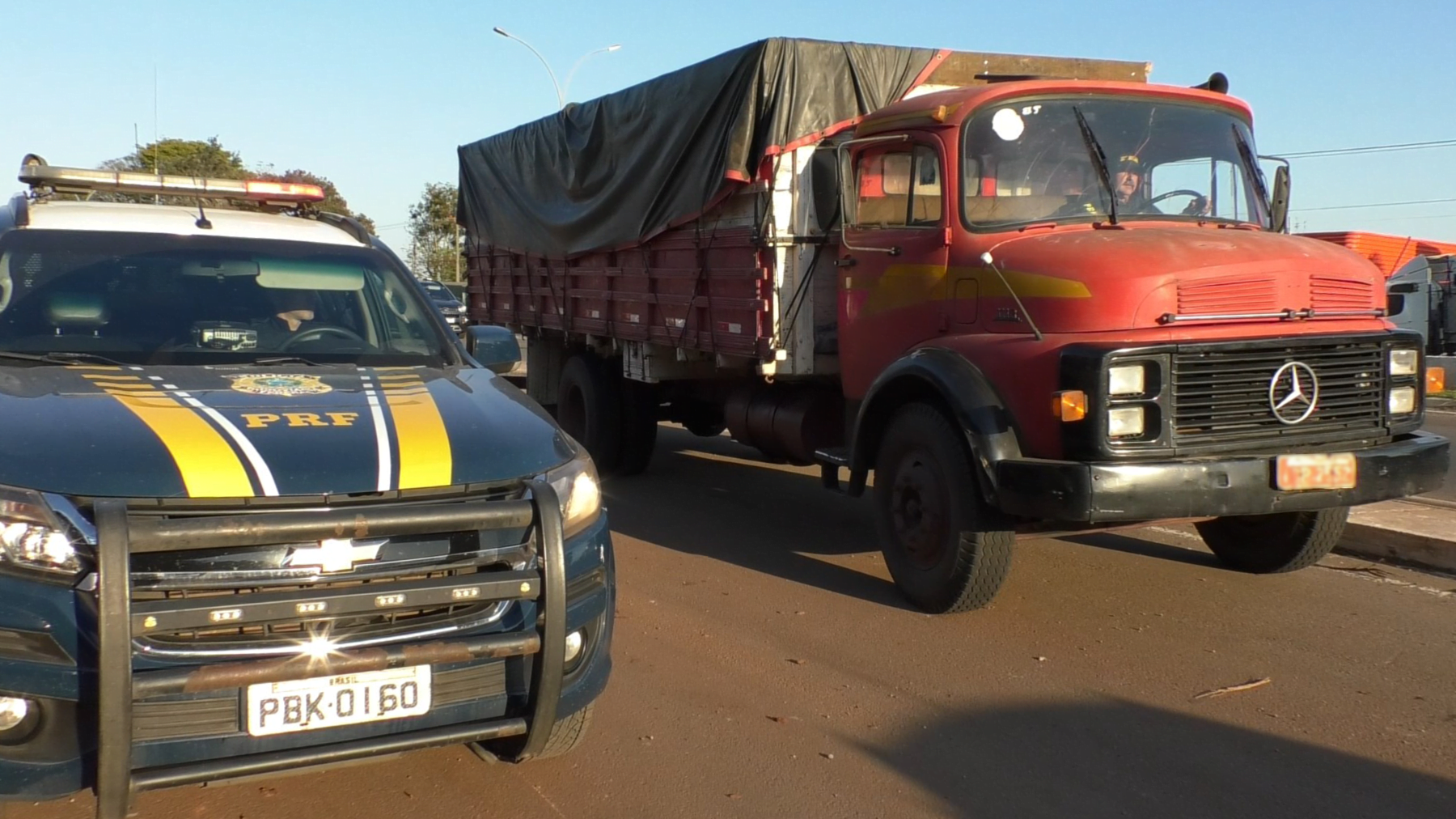 Foto de viatura da Polícia Rodoviária Federal ao lado de caminhão vermelho.