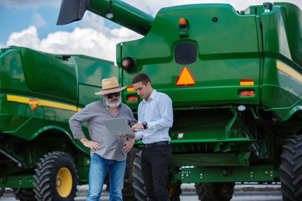 Foto de homem com roupa social e agricultor conversando perto de máquina agrícola.