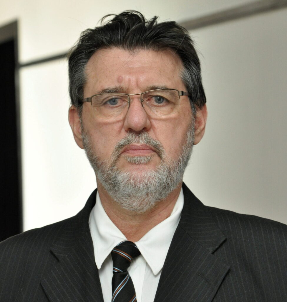 A foto mostra um homem de meia idade com cabelo escuro e barba grisalha. Usando terno e gravata.