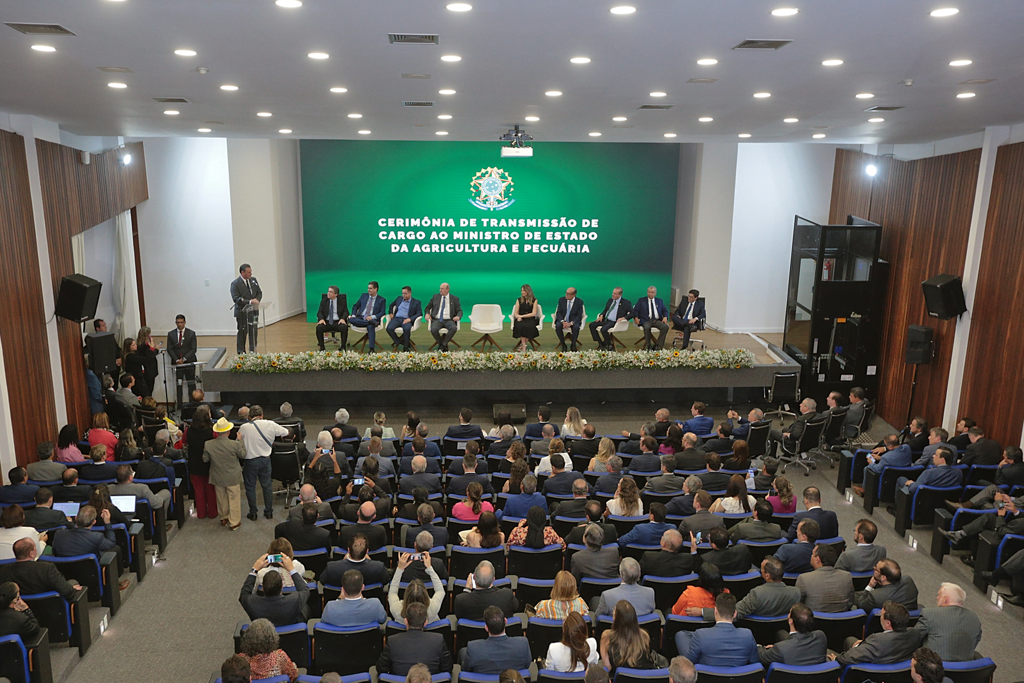 Foto de pessoas em auditório enquanto Carlos Fávaro discursa no palco. Ao fundo, um telão exibe a frase: Cerimônia de Transmissão de cargo ao ministro de Estado de Agricultura e Pecuária.