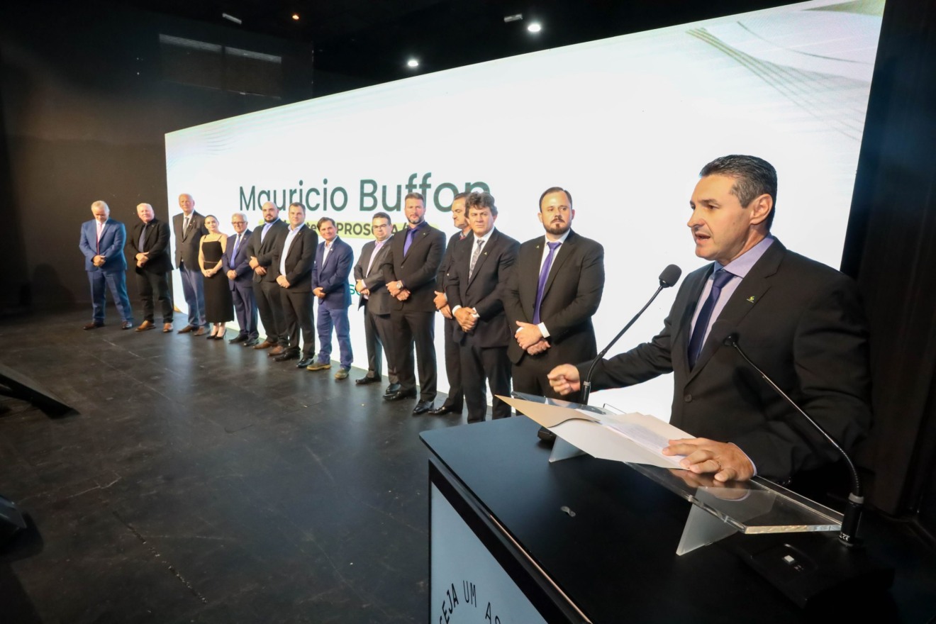 Foto de Maurício Buffon discursando em palco. Atrás dele está uma fila com vários homens e uma mulher.