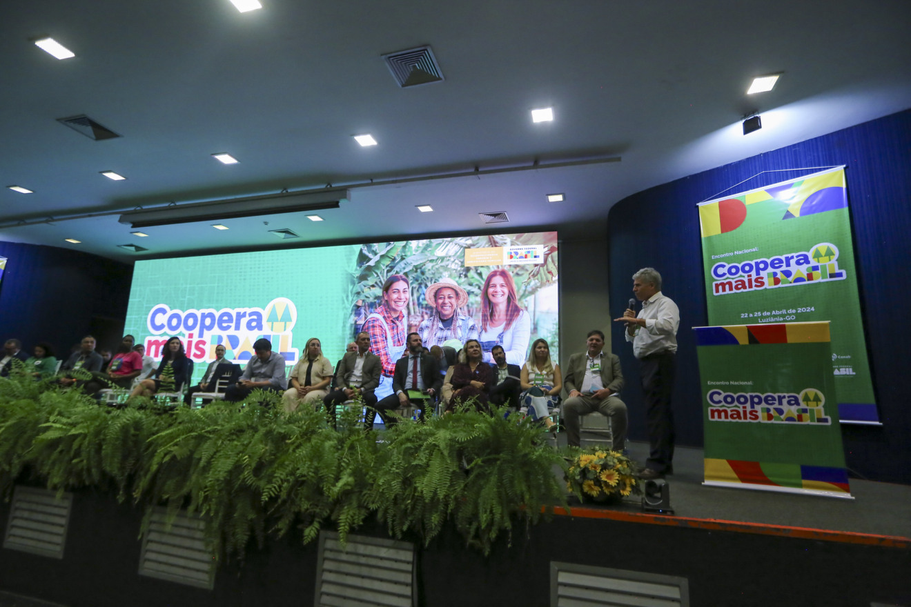 Foto de palco com várias pessoas sentadas enquanto o Ministro Paulo Teixeira, em pé, discursa. O telão no fundo e banners anunciam o 