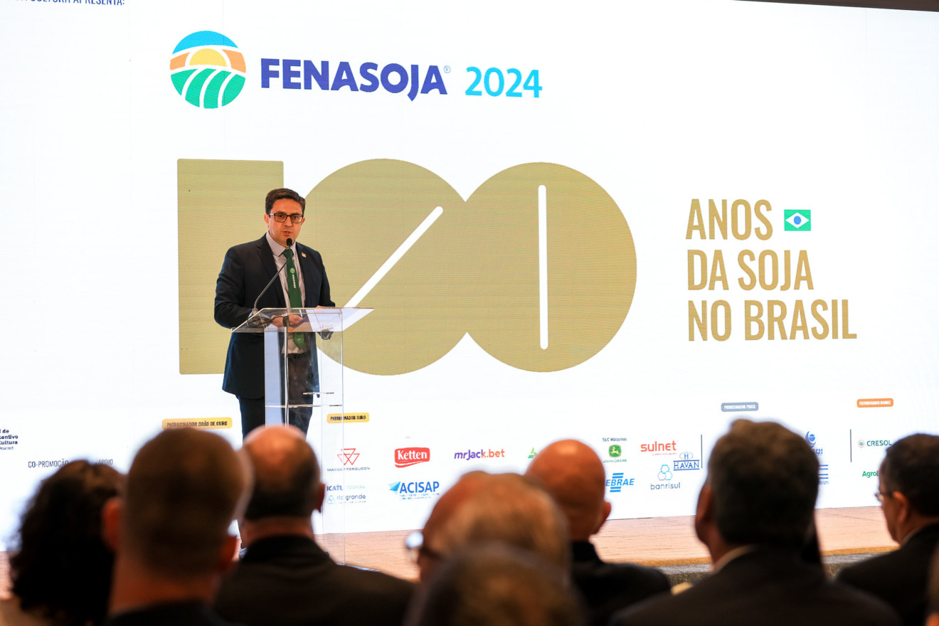 Foto de homem, com terno e gravata, falando em parlatório em um palco. Atrás dele está um telão anunciando a Fenasoja 2024 - 100 Anos da Soja no Brasil.