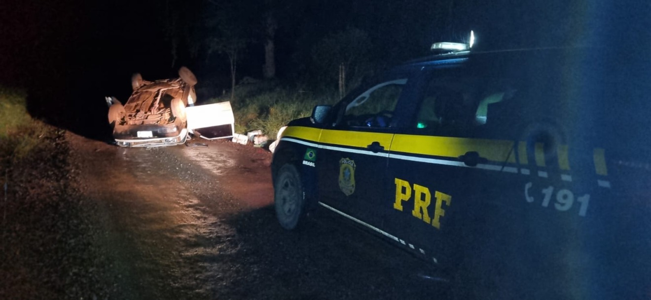 Foto viatura da PRF próxima a carro capotado com galões de agrotóxicos caídos no chão.