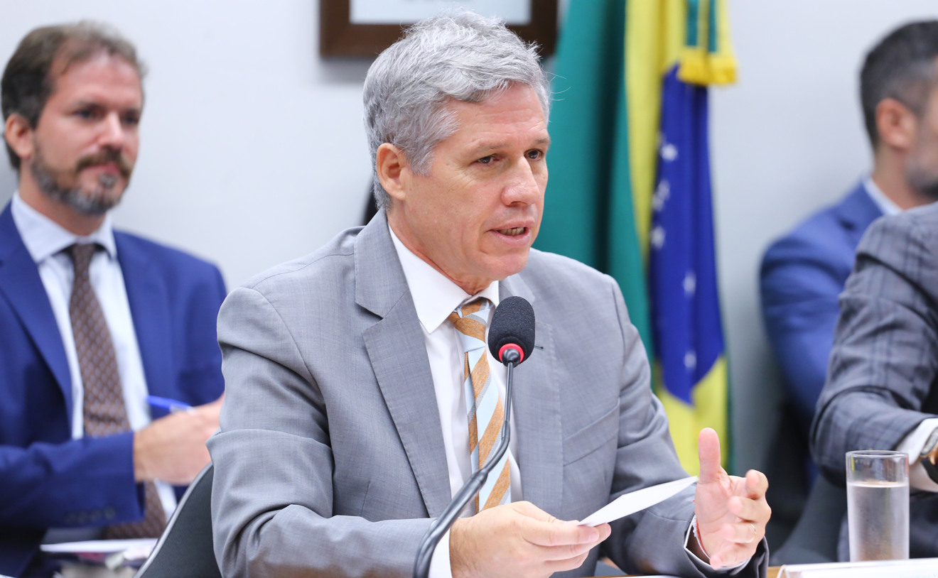 Foto de ministro Paulo Teixeira falando em microfone. Ele é branco, tem cabelo grisalho e usa terno cinza com gravata cinza e laranja.