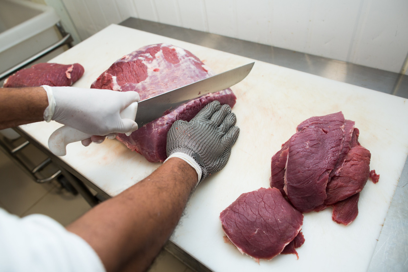 Foto focada em mãos com luvas cortando carne com faca.