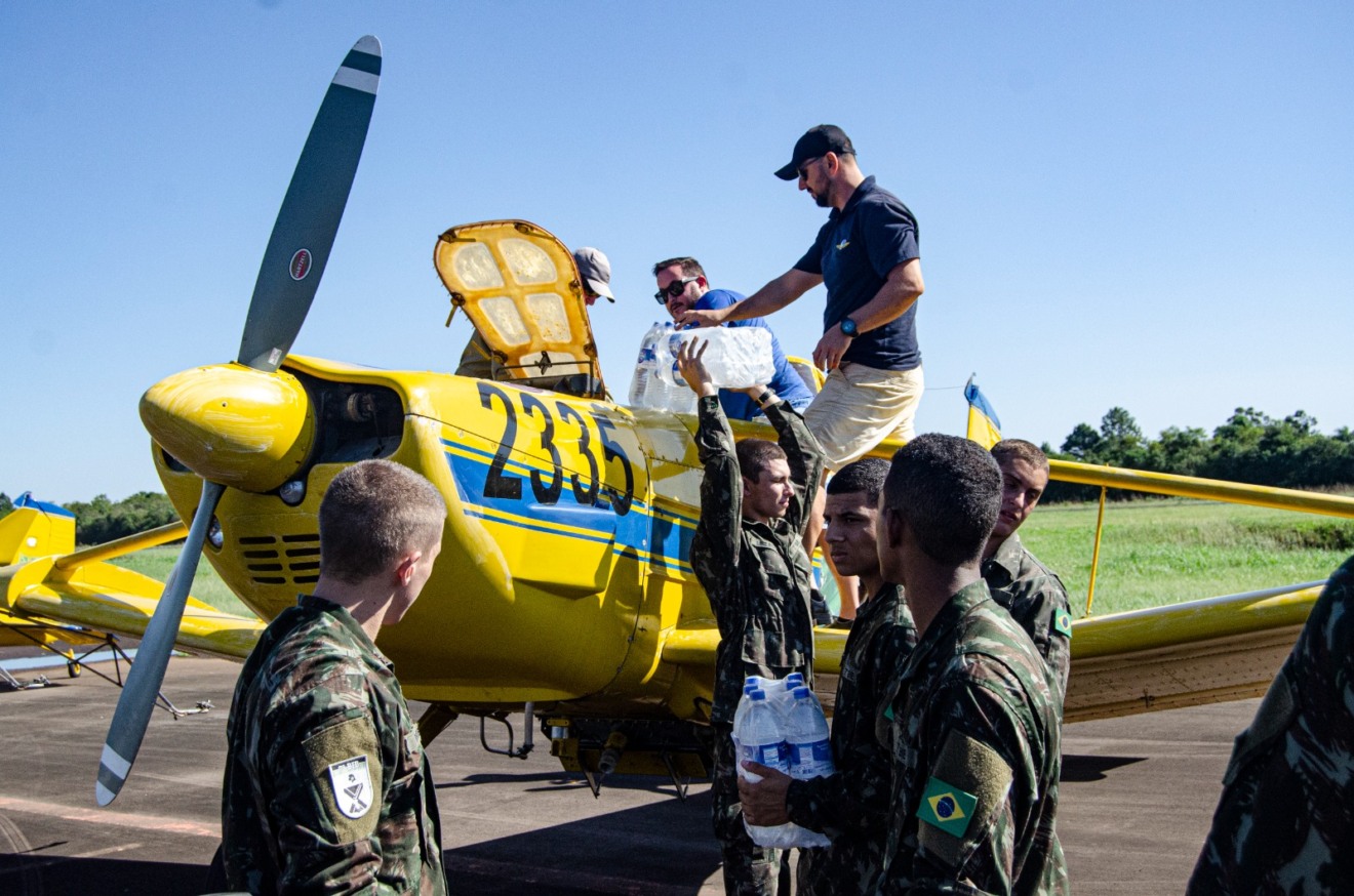 Foto de um grupo de militares alcançando água para dois homens estão colocando as garrafas em um avião agrícola amarelo.