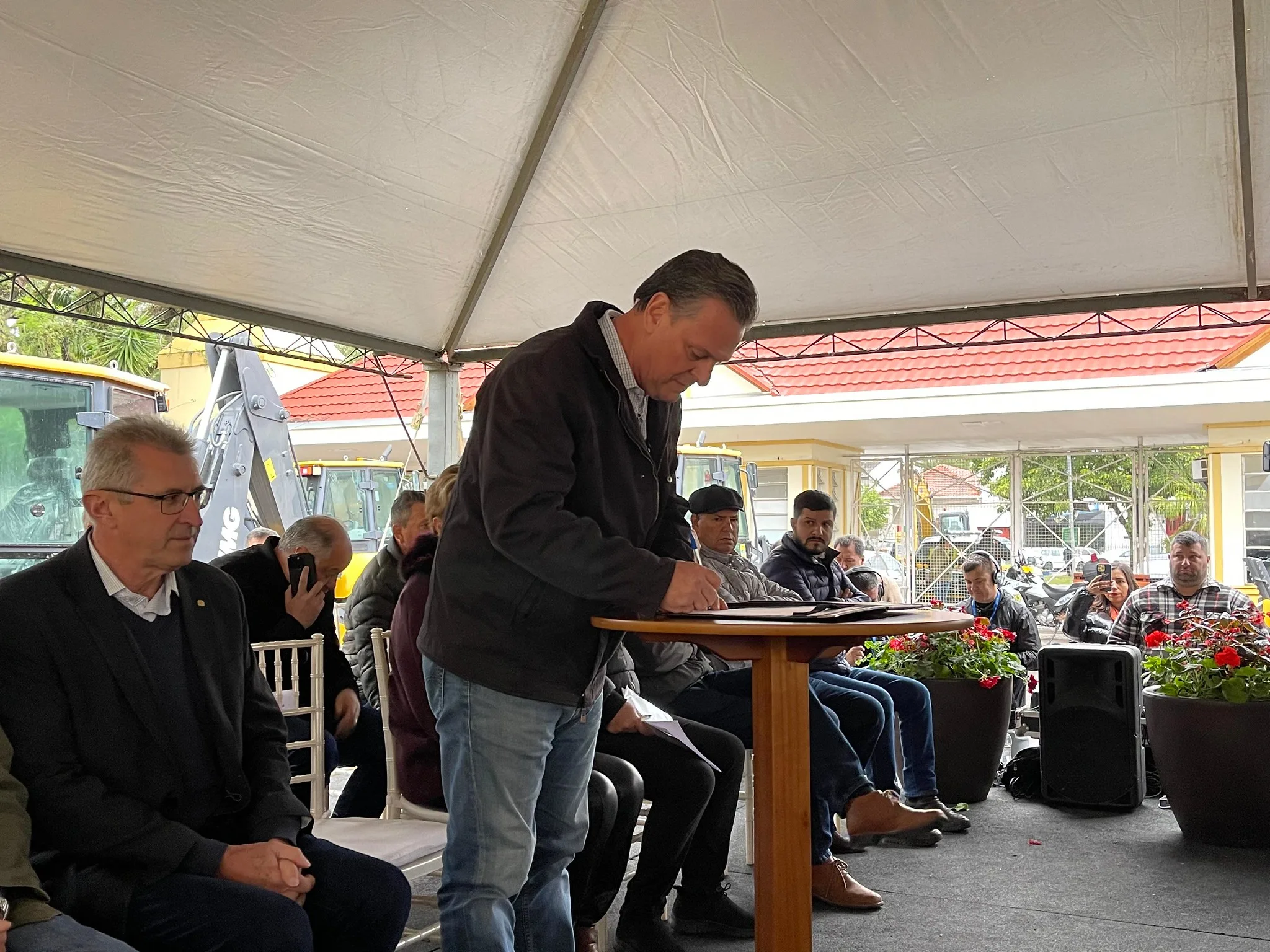 Foto de ministro Carlos Fávaro em pé enquanto assina papéis que estão sobre uma mesa. Ele está embaixo de uma lona de plástico e várias pessoas estão sentadas atrás dele.