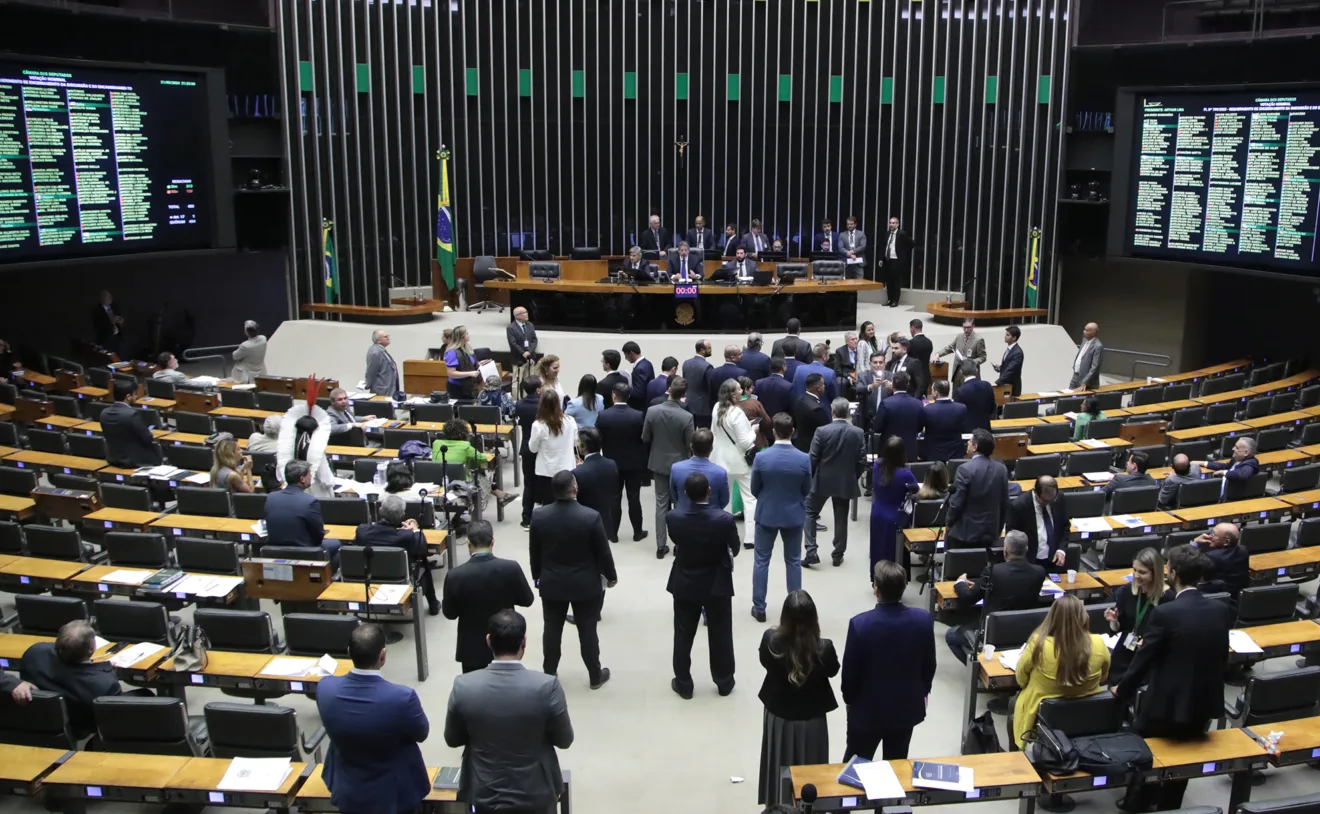 Foto de deputados no plenário da Câmara de Deputados durante Sessão Deliberativa. A maioria está em pé em frente a mesa da presidência.