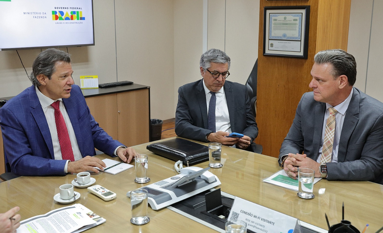 Foto dos ministros Carlos Fávaro e Alexandre Padilha conversando ao redor de mesa.