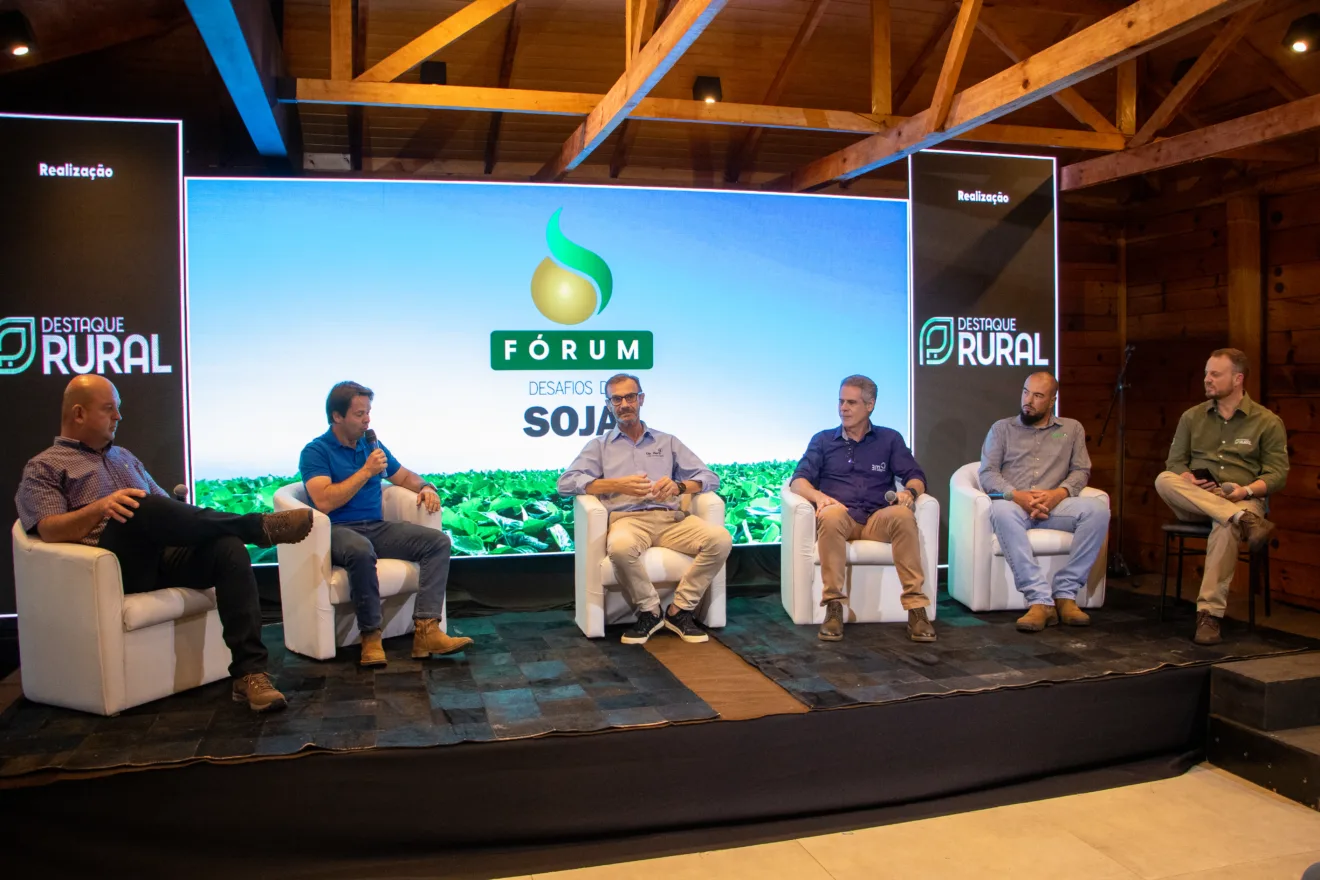 Foto de cinco homens sentados em poltronas em frente à telão com logomarca do Fórum Desafios da Soja. Um deles segura um microfone.