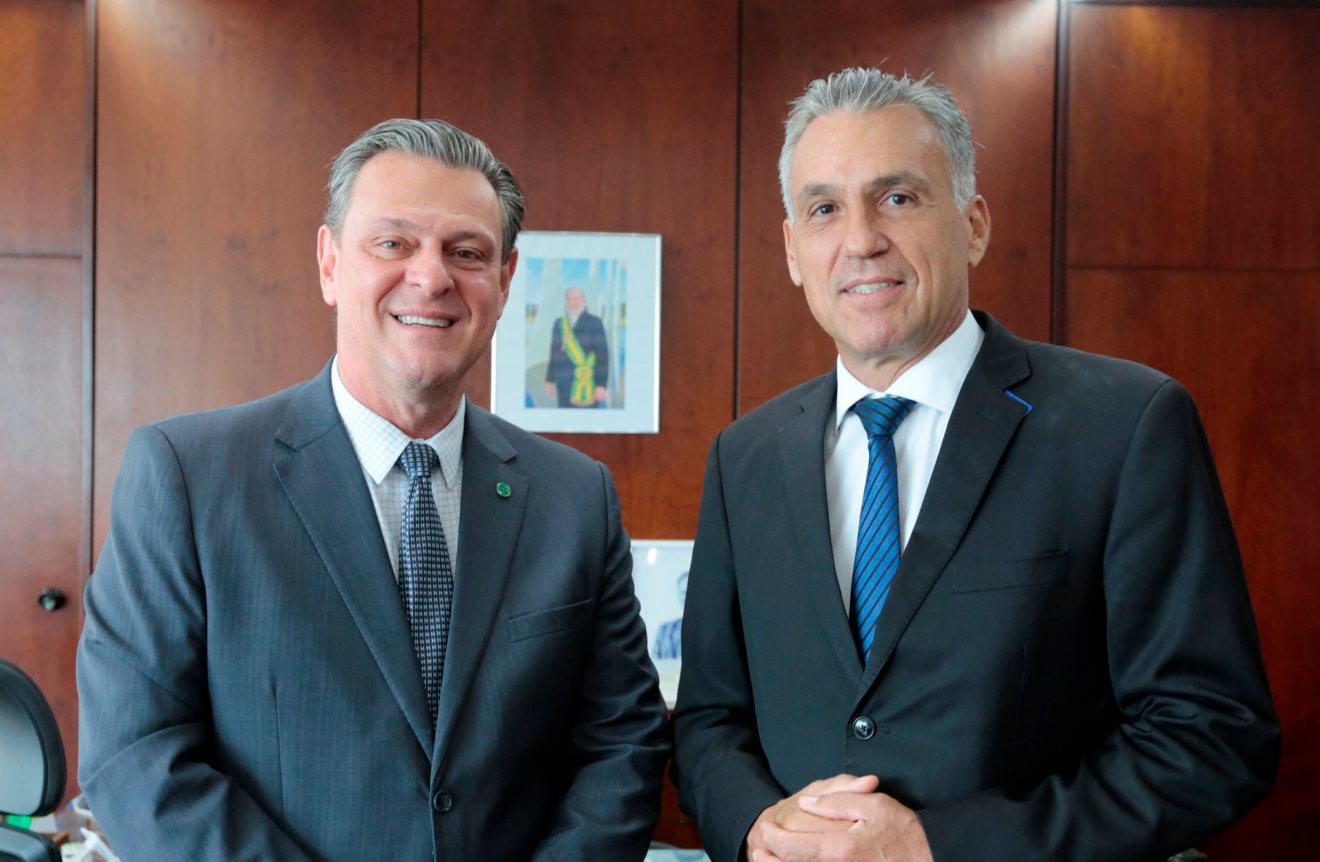 Foto de dois homens sorrindo enquanto posam para foto. Ambos são brancos, tem cabelo grisalho e usam ternos.