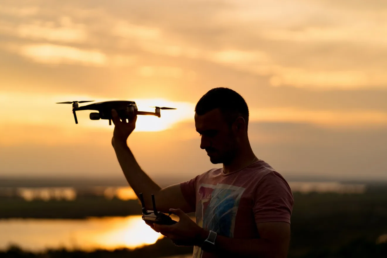 Silhueta do homem pilotando um drone ao pôr do sol com céu ensolarado no fundo.