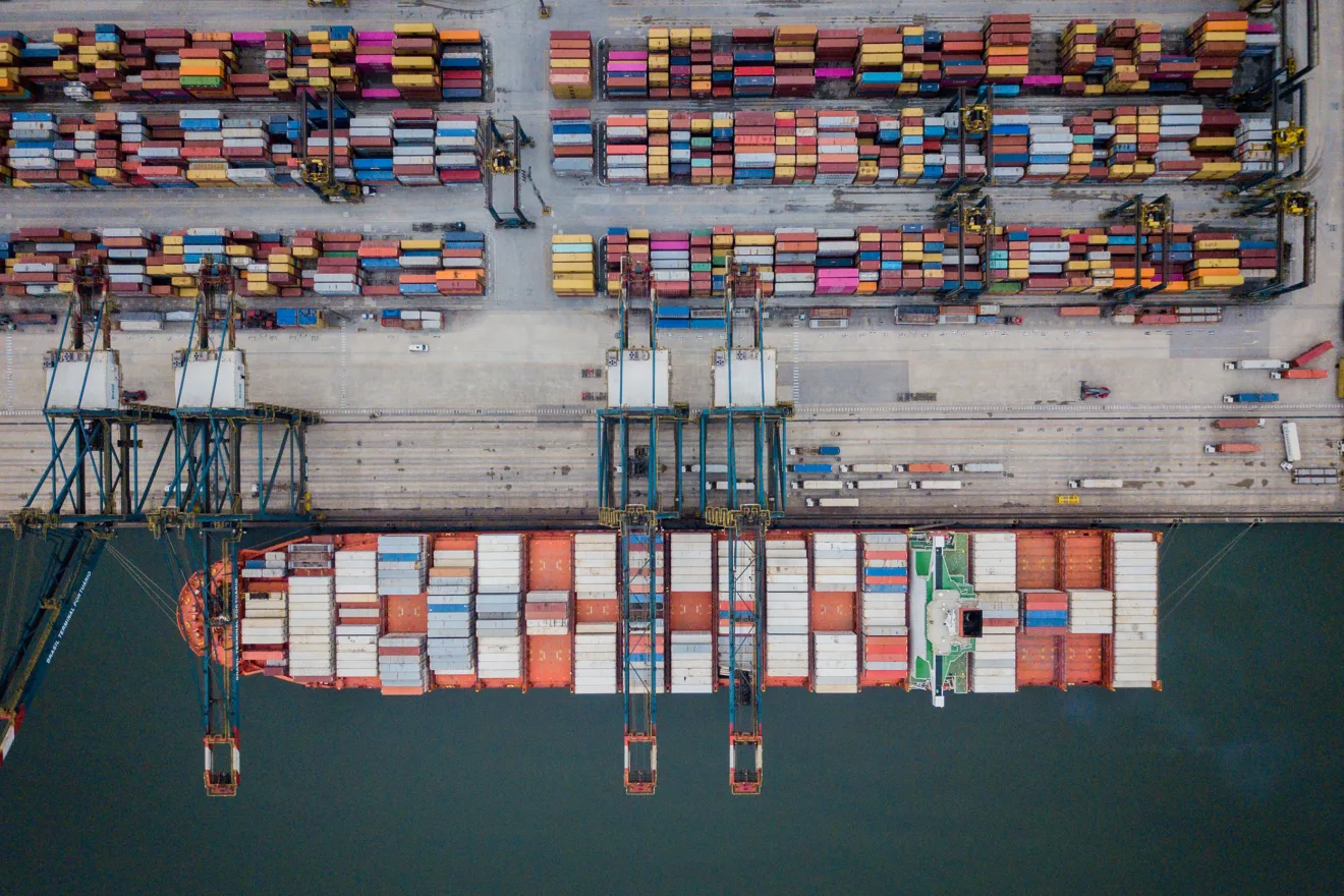 Foto de navio com containers ao lado de em porto onde estão mais containers.