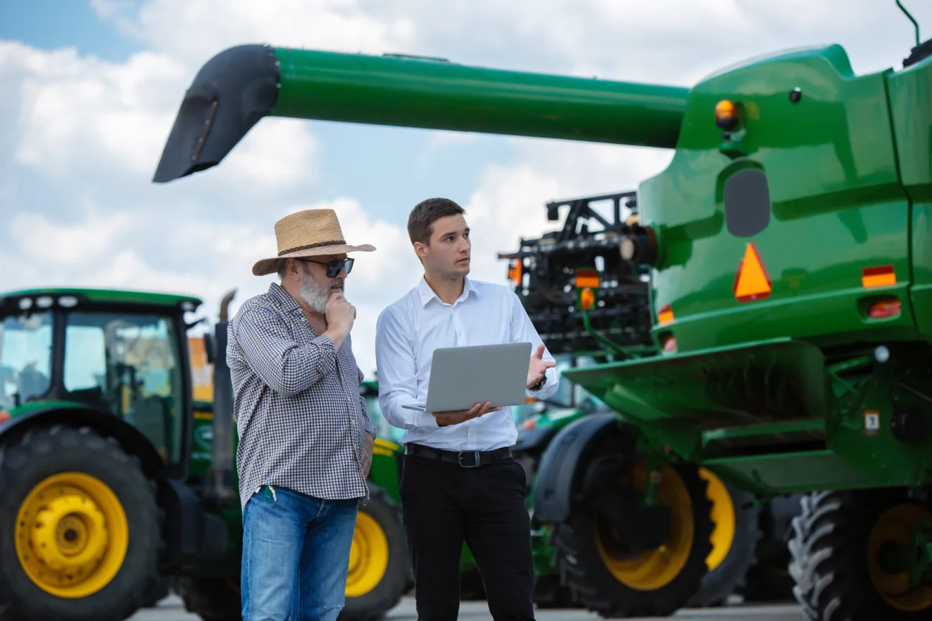Foto de dois homens ao lado de máquina agrícola verde olhando para o notebook que um deles segura.