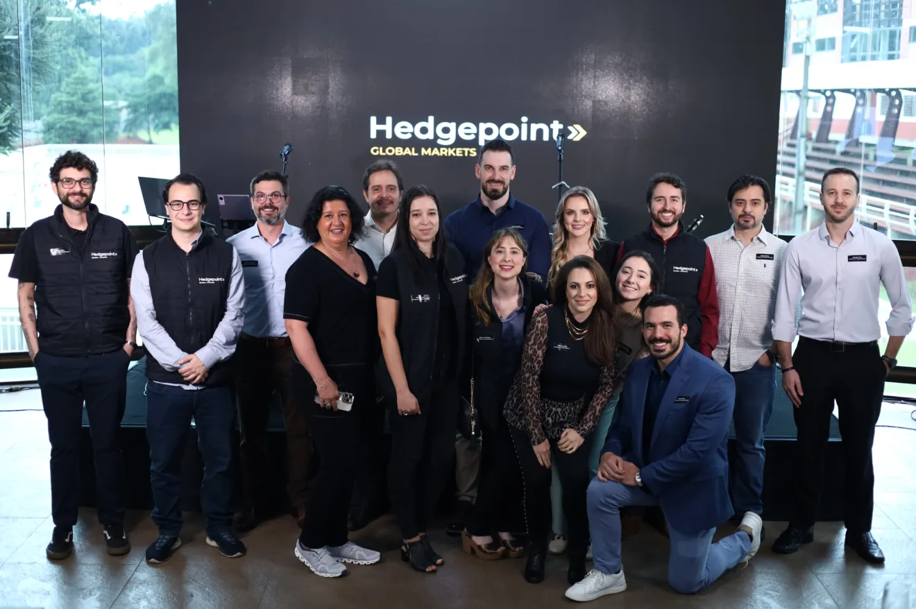 Foto de pessoas posando para foto em frente a letreiro com a logo da Hedgepoint Global Markets. Elas estão sorrindo para foto e a maioria usa roupas escuras.
