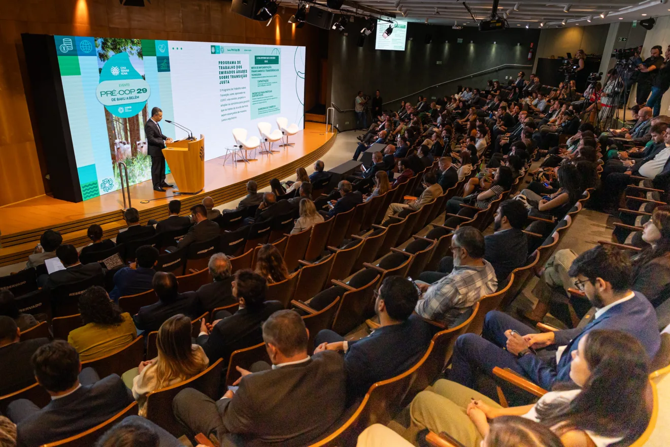 Foto de auditório com pessoas sentadas assistindo enquanto homem de terno fala em parlatório no palco em frente a um telão.