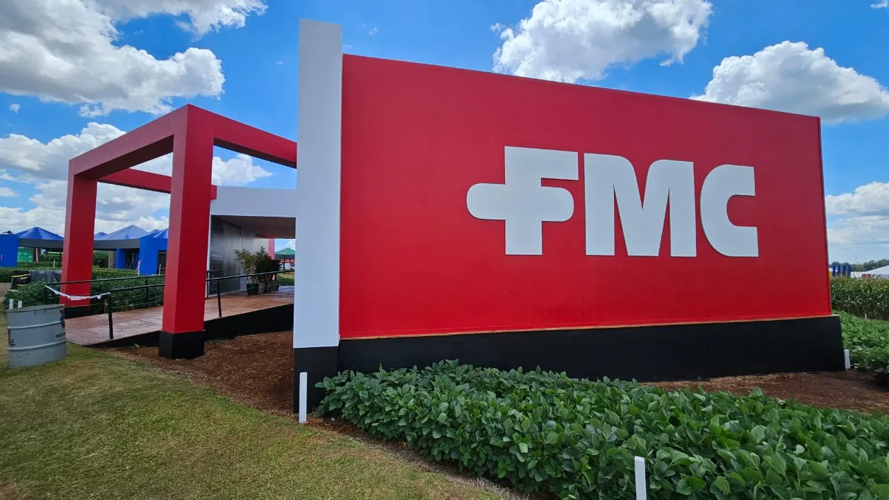 Foto de estande da FMC. Ele tem um painel grande vermelho com FMC escrito em branco. Na frente há plantas de soja e ao fundo está a entrada do estande.
