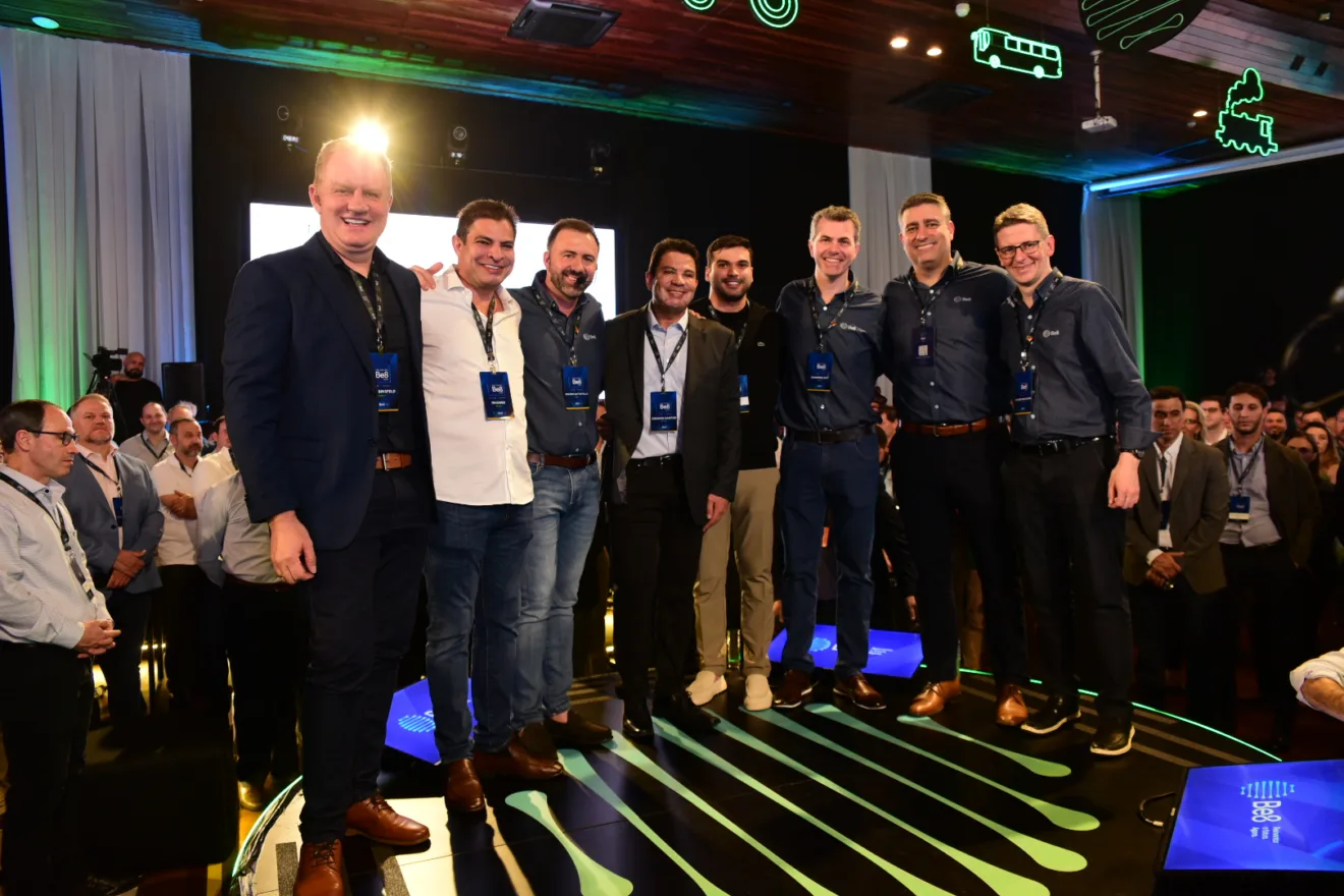 Foto com oito homens, lado a lado, com roupas formais durante evento. Eles sorriem para a foto.