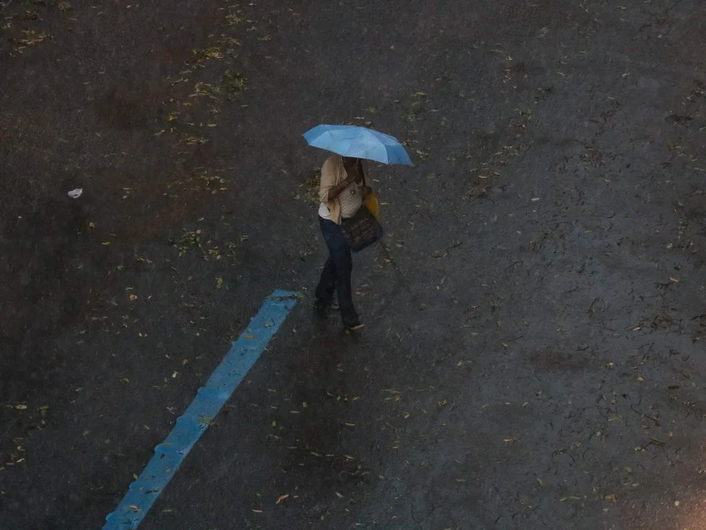 Foto de pessoa, em andando na rua com guarda-chuva enquanto chove.