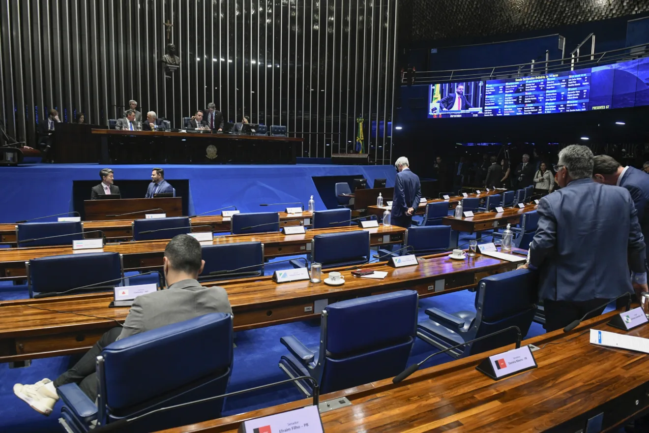 Foto do Plenário do Senado Federal. Em primeiro plano estão as mesas com pessoas sentadas e em pé atrás delas. No fundo está a mesa diretora do Senado e painéis.