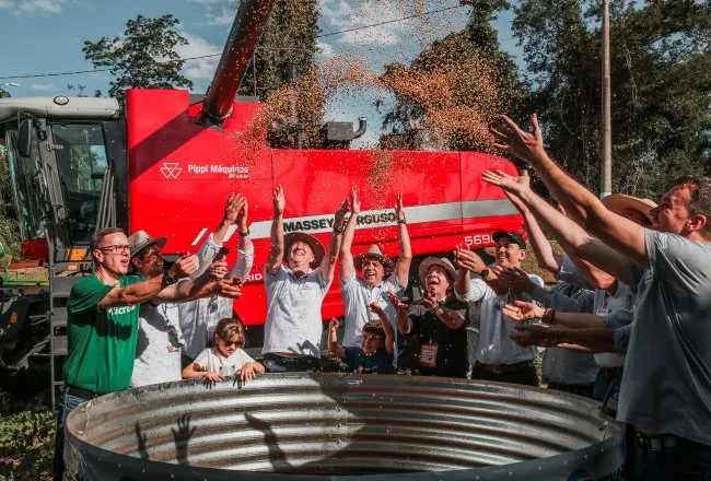 Foto de pessoas ao redor de tonel com grãos de soja caindo. Atrás deles está uma máquina agrícola vermelha.